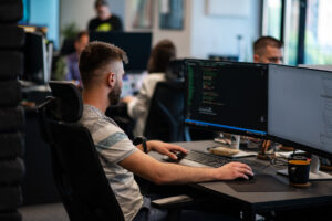 Jeune expert en informatique travaillant dans son bureau de coworking moderne et récent. Le programmeur est entrain de coder sur son ordinateur. L'homme est assis à son bureau dans un espace partagé, avec des collègues en arrière-plan.